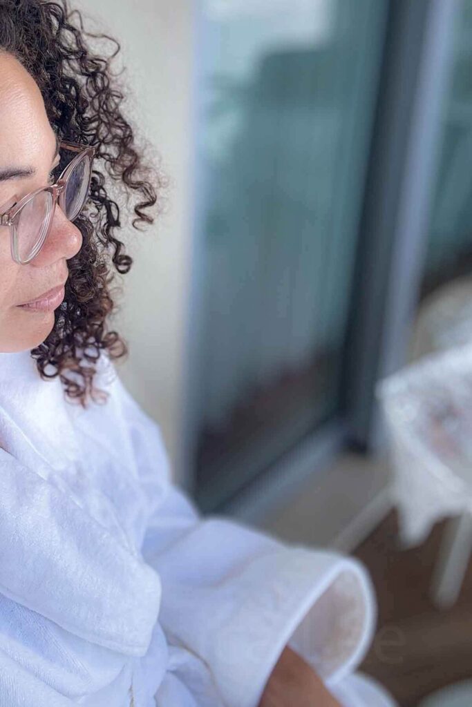 Side profile of a woman with curly brown hair and glasses hair in a white robe enjoying a quiet moment on a hotel balcony in Laguna Beach.