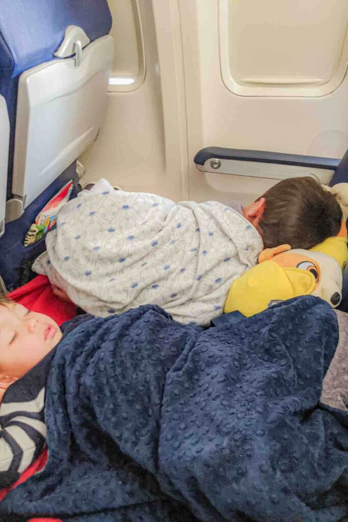 Two toddlers sleeping across airplane seats with inflatable footrests for more space, blankets, and a stuffie pillow during a long distance flight. A cozy setup makes it easier when flying long haul with toddler overnight.