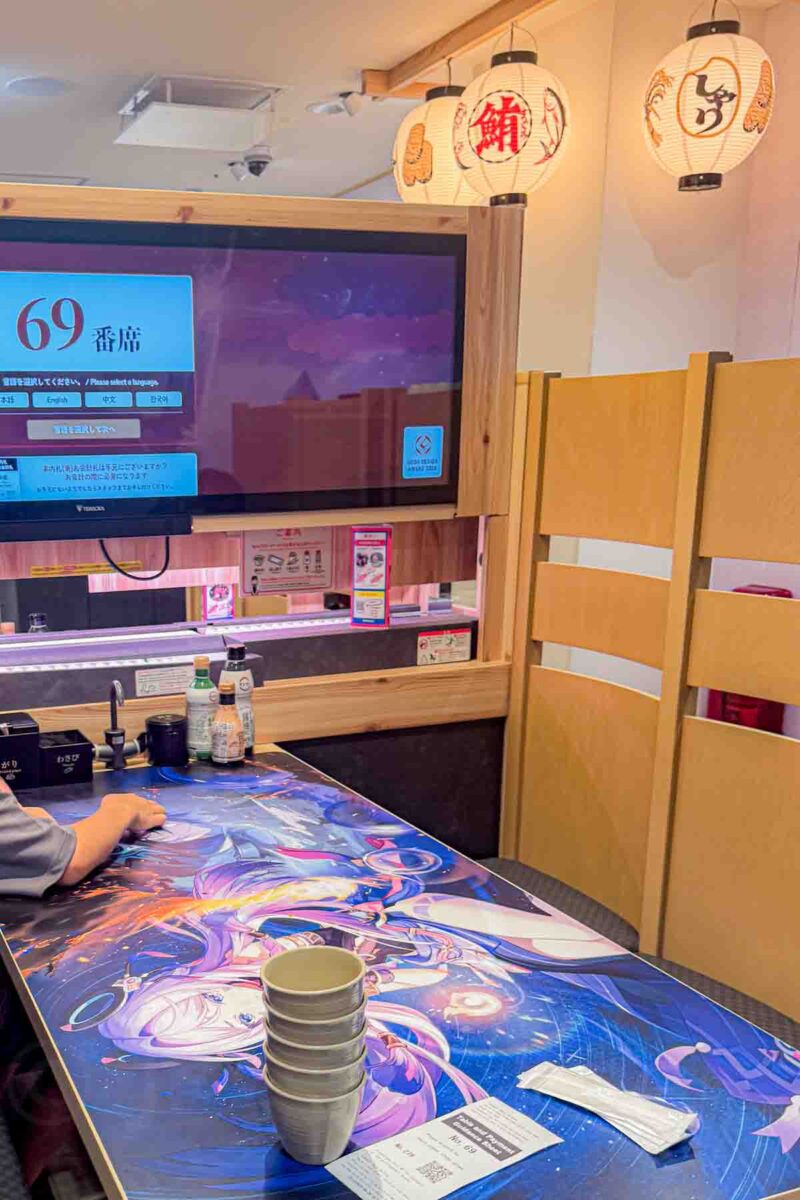 Restaurant booth at Sushiro in Osaka with a bright anime themed table and a digital ordering screen displaying “69 番席.” A playful, interactive dining setup that keeps kids engaged while eating in Japan.