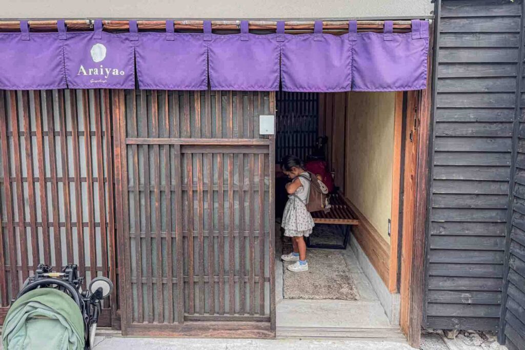 A child buckles their backpack while standing at the entrance of a traditional Tokyo townhouse with wooden sliding doors and purple noren. Example of family friendly lodging when deciding where to stay in Tokyo with kids.