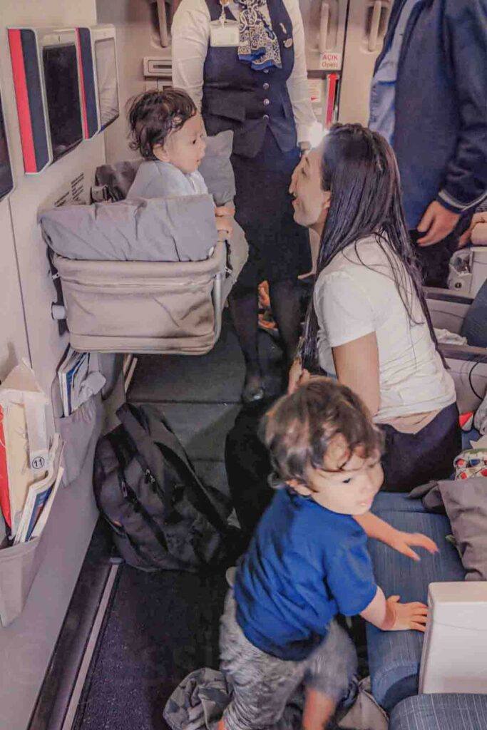 A mother sits across and interacts with a smiling baby sitting upright in an airplane bassinet while a flight attendant stands nearby during boarding. The cabin moment shows how airlines support families flying long haul with babies.