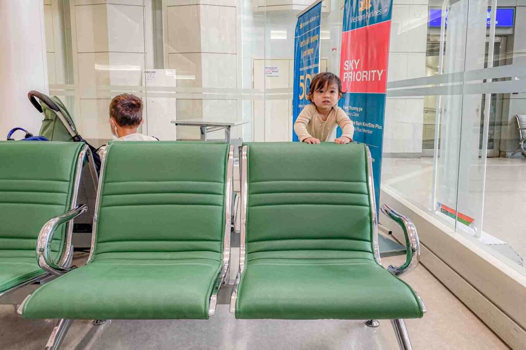 A baby peeks over rows of green seats beside a stroller at the boarding gate in Taipei Taoyuan Airport. Another child faces the other way busy with an activity next to a stroller. The quiet waiting moment shows traveling with multiple kids at the airport before an international flight.