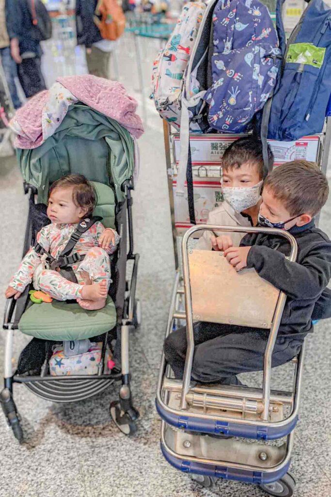 A baby sits in a yoyo stroller while two brothers ride a luggage cart stacked with backpacks in a busy international terminal. The scene captures family travel logistics with several young children.