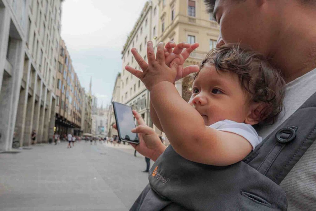 Baby riding in a front carrier reaches both hands into the air while a parent looks at their phone trying to figure out directions in Italy. The baby looks curious and engaged while being carried comfortably on the go.
