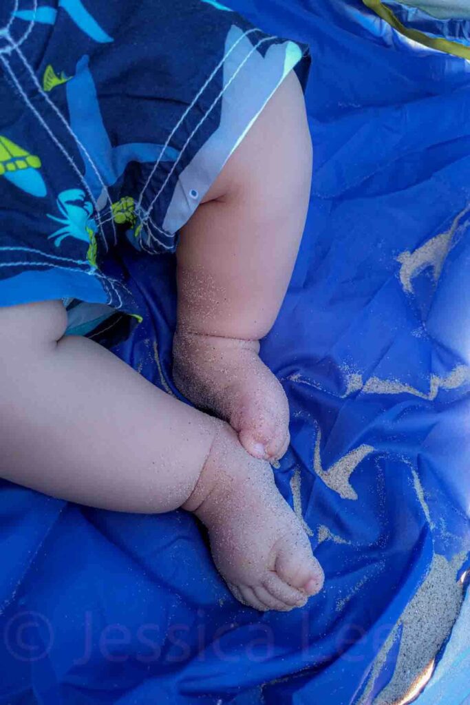 Close up of chubby baby legs in blue swim shorts resting on a bright blue beach mat with sand clinging to tiny toes. The relaxed posture suggests a calm beach day with minimal fuss.