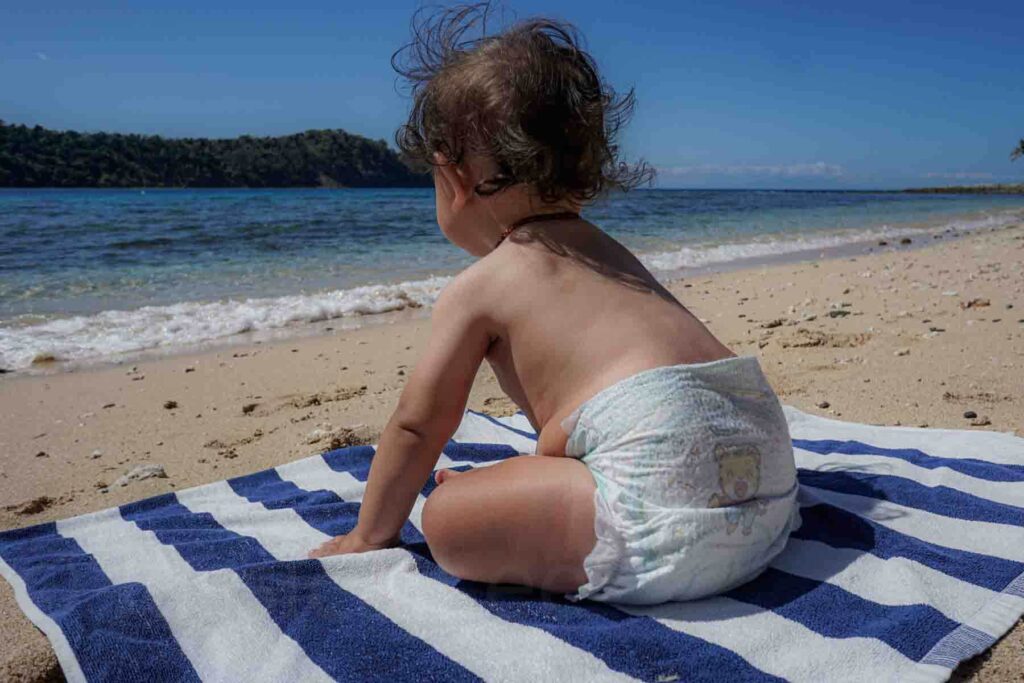 Baby only wearing a diaper sits on a blue and white striped towel on the sand facing the ocean with gentle waves rolling onto a sandy beach on a private resort island in Fiji. The child leans forward checking out the sand while looking out at the water. Scenes like this show why many parents consider this the best age to travel with a baby for relaxed beach trips overseas.