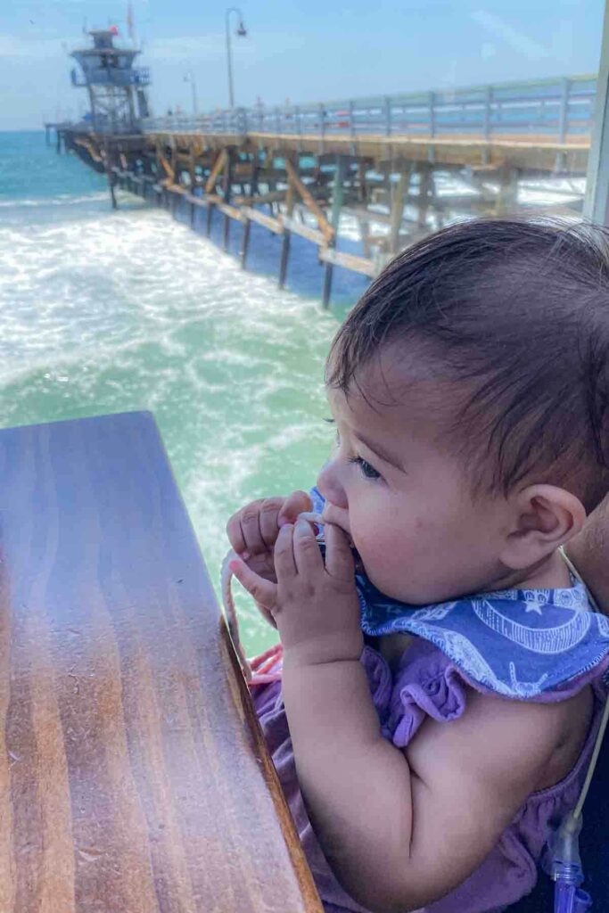 Baby sits at a wooden table at a pier restaurant overlooking the ocean beside a long pier stretching over the water in San Clemente, California. Waves crash below as the child gnaws on a teether.