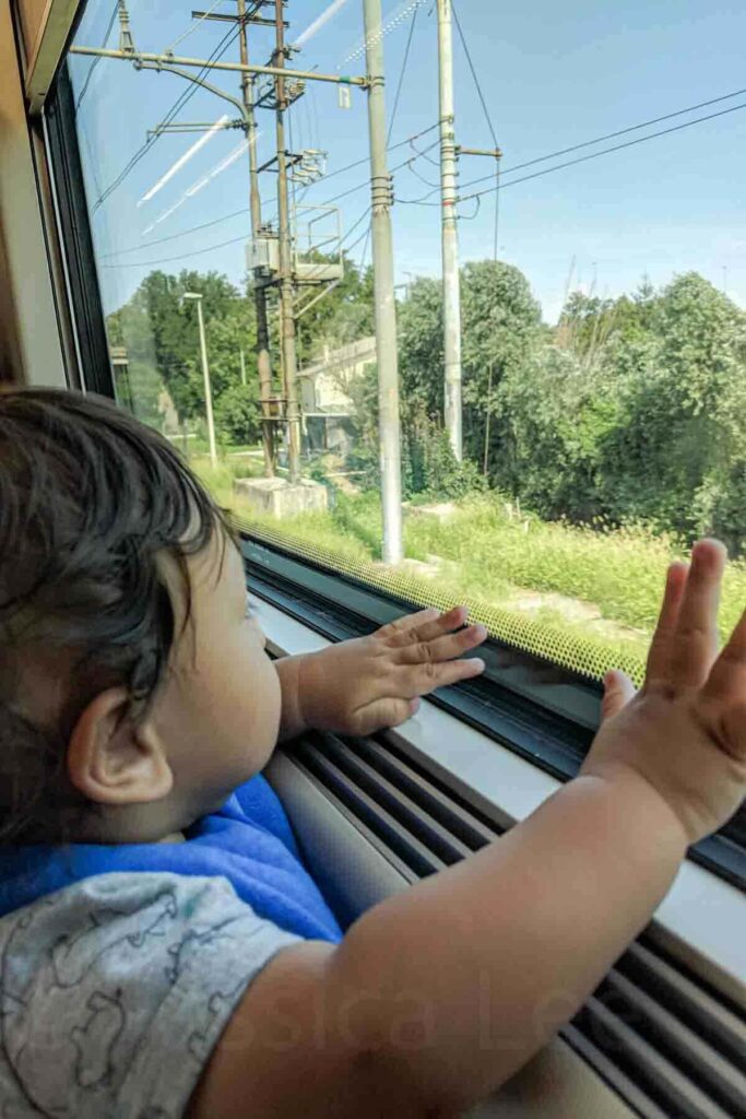 Baby presses both hands against a high-speed train window while watching green Italian countryside blur past outside. The child appears fascinated by the movement and changing scenery. Younger babies curiosity makes simple transit part of the adventure.