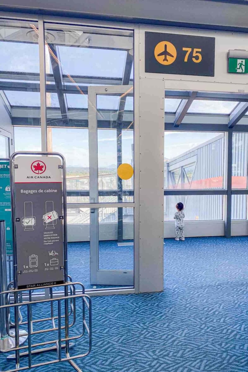 Toddler stands by airport windows near gate 75 looking outside at the runway while waiting for a long flight, an Air Canada cabin baggage sizer stands in the foreground.