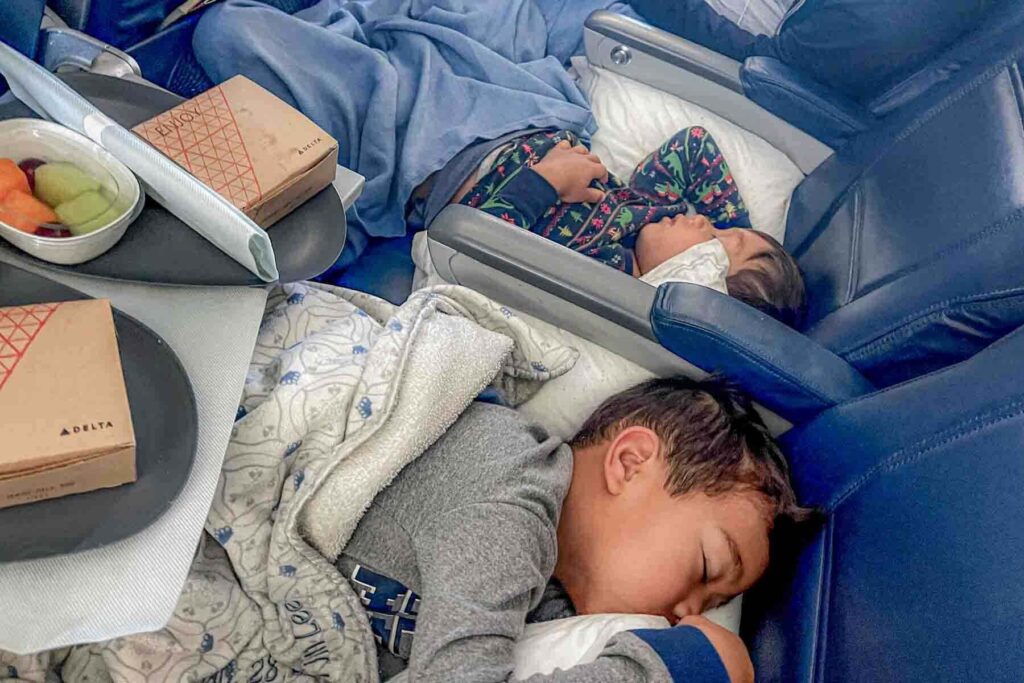 Two brothers sleep across airplane seats under blankets with open tray tables with fruit and airline snack boxes labeled "Delta". The quiet cabin scene shows siblings resting on an overnight flight.