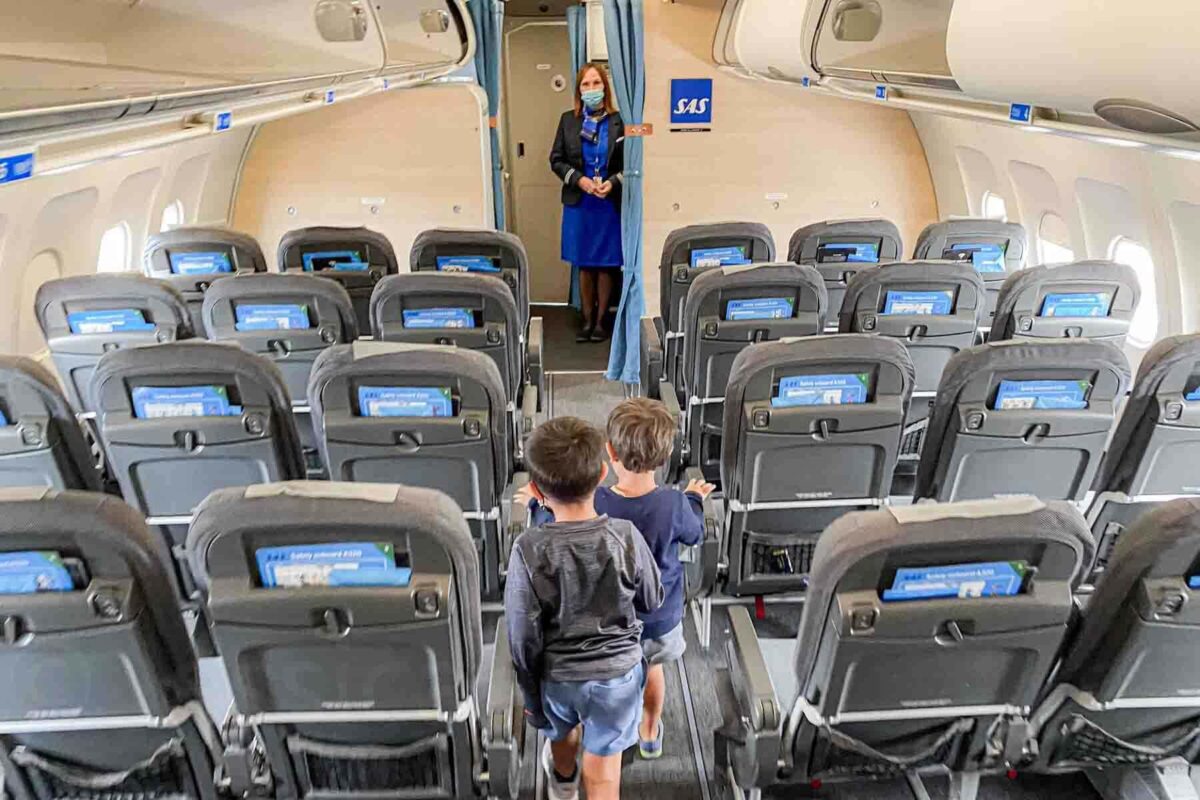 Two boys walk toward a flight attendant on a SAS airplane with a bright blue branded sign at the front near the exit after flying.