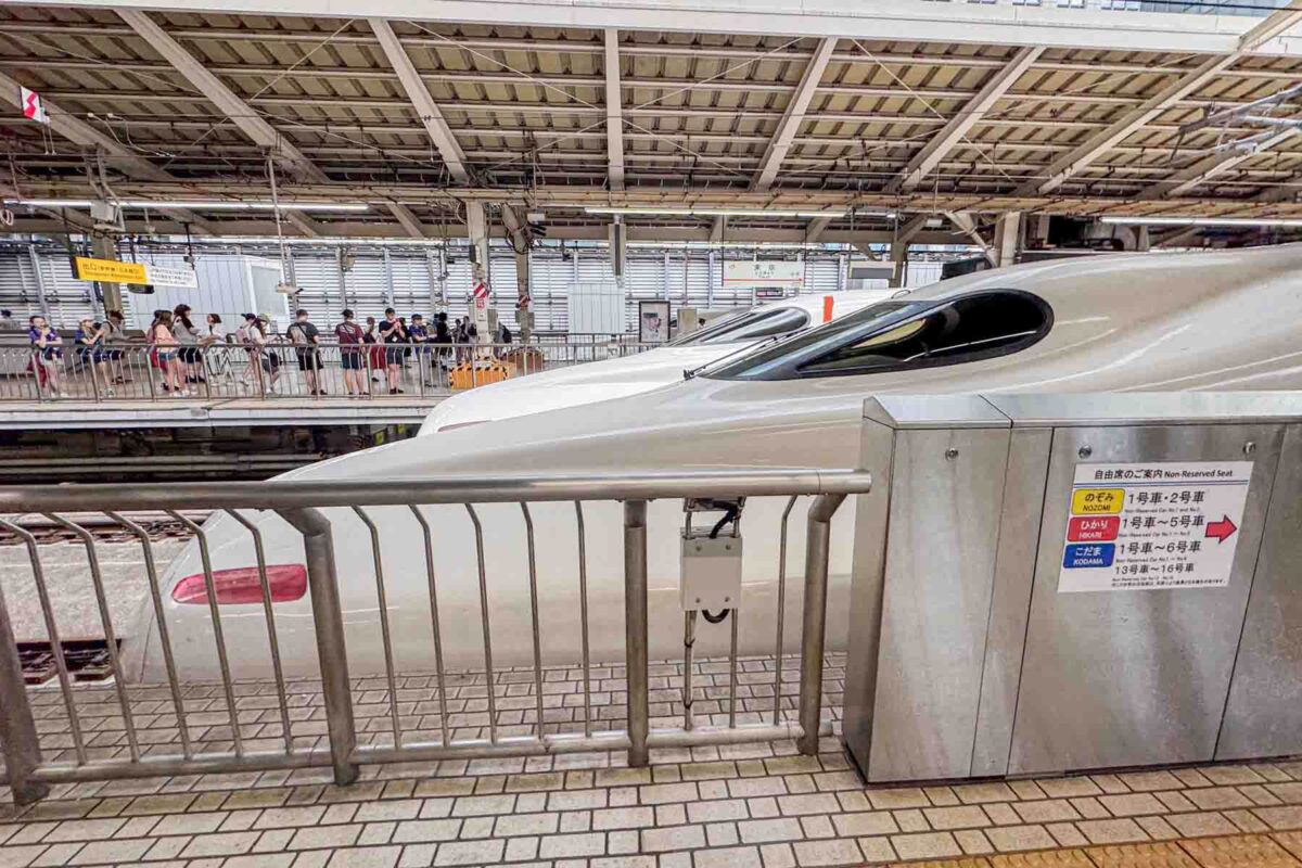 Sleek Shinkansen bullet train nose at a busy station platform in Japan with passengers waiting behind railings. Shows how Japan train travel with kids can be fast efficient and visually exciting.