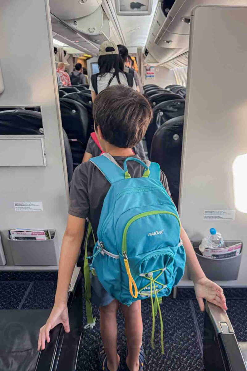 Young child with a bright blue backpack walking down the aisle of an airplane after landing in Okinawa. Highlights domestic flights as part of getting around Japan with kids between cities.