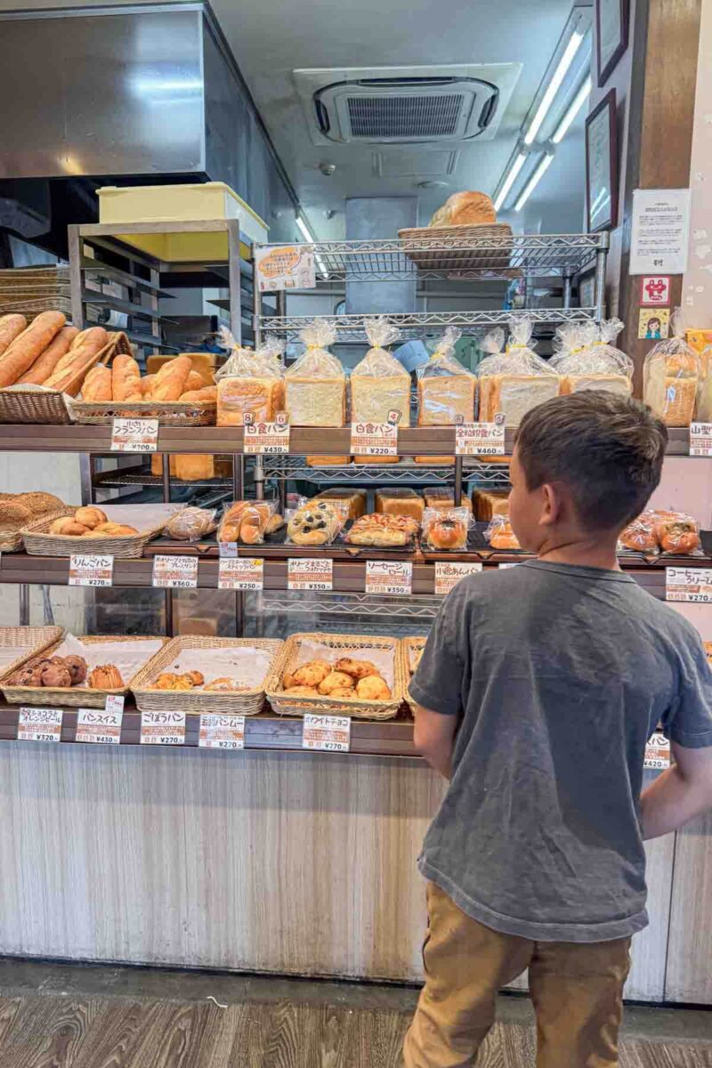 Child standing at a Japanese bakery counter in Setagaya City in Tokyo looking at neatly packaged breads and pastries with price labels in yen. A glimpse of everyday stops while exploring neighborhoods during a family trip to Japan.