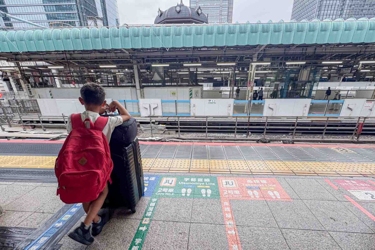 Child with a red backpack leaning on a large suitcase while waiting on a marked train platform in Japan. Platform signs and lines show how to navigate trains around Japan with kids.
