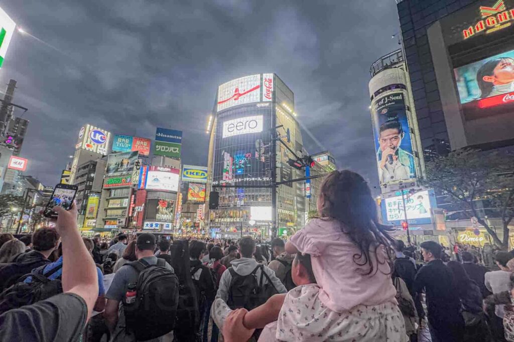 Child wearing a pink t-shirt sitting on a parent’s shoulders watching crowds cross Shibuya Crossing at night surrounded by bright neon lights in central Tokyo.