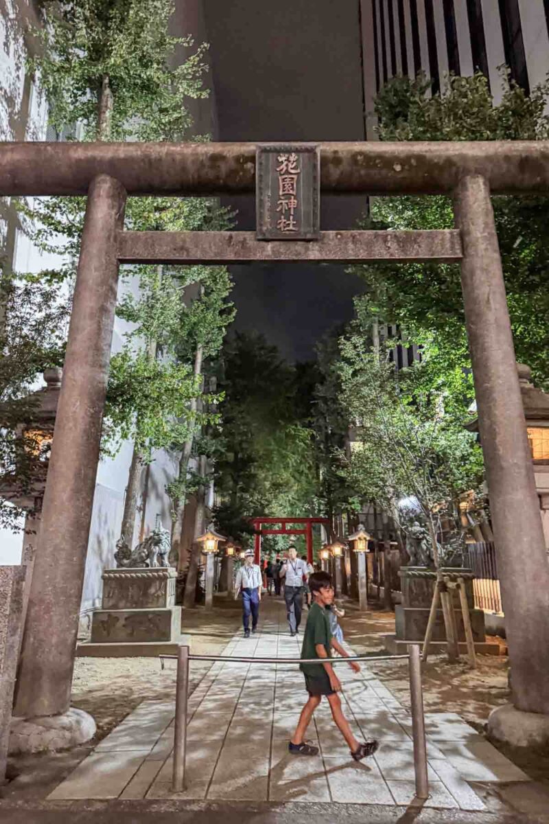 Child walking beneath a large stone torii gate at the Homotsuden shrine entrance at nighttime in Tokyo surrounded by trees and lanterns. Shows cultural stops easily reached while walking around Japan with kids.