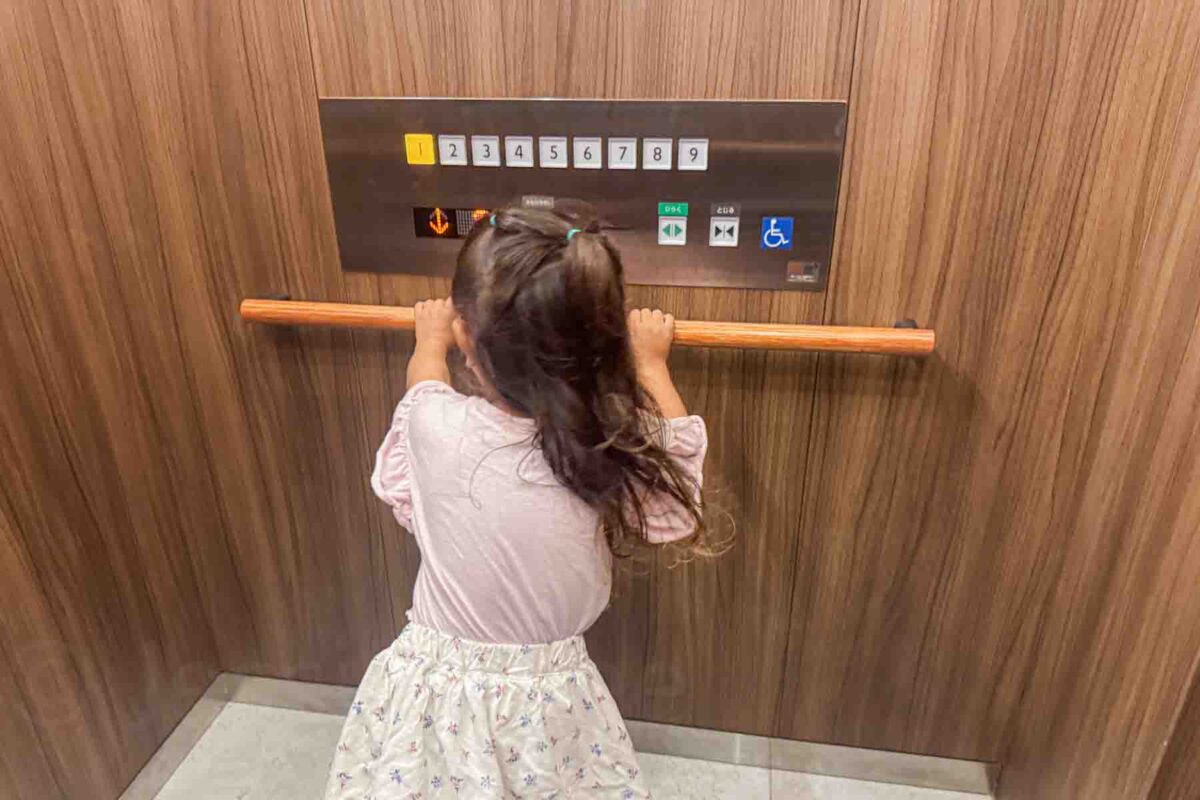 Young child standing on tiptoes in a hotel elevator with buttons labeled "1 2 3 4 5 6 7 8 9" inside a wood paneled lift. Highlights accessibility features useful for Japan stroller travel.
