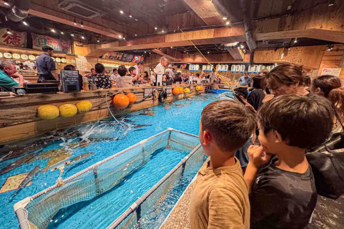 Indoor fishing pool inside a restaurant surrounded by diners as children watch fish swim beneath the surface. A unique entertaining dining concept for kids in Japan.