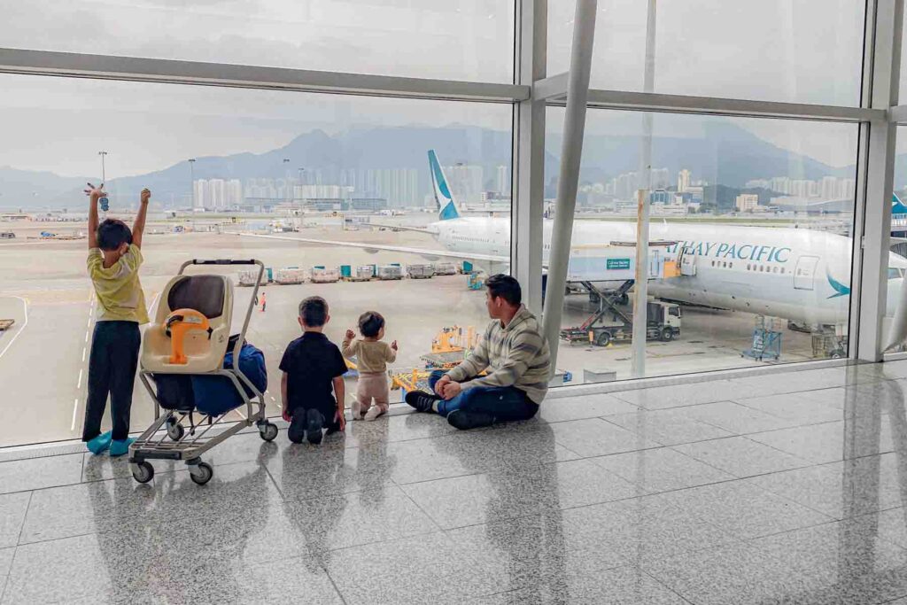 A dad sits on the airport floor with three children looking through large windows at a Cathay Pacific airplane outside in Hong Kong airport. The family waits together before boarding their long international flight.
