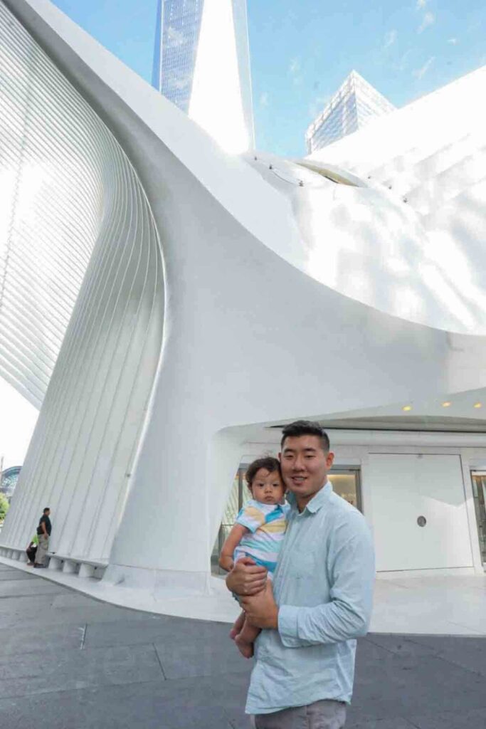 Parent holds baby in front of the white curved architecture of the World Trade Center in New York City. The modern structure towers overhead while the baby rests comfortably in their arms. Babies are often content with city sightseeing while traveling.