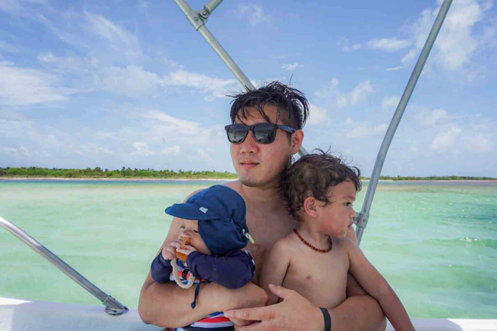 Father holds two young children on a boat surrounded by calm turquoise water off Playa del Carmen in Mexico and a distant shoreline. The baby wears a navy sun hat while the toddler looks out over the water.