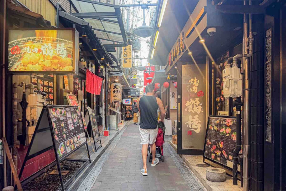 Parent pushing a stroller down a narrow restaurant lined alley in Osaka with large picture menus and lantern signs on both sides. Shows real conditions for Japan stroller travel in tight urban spaces.
