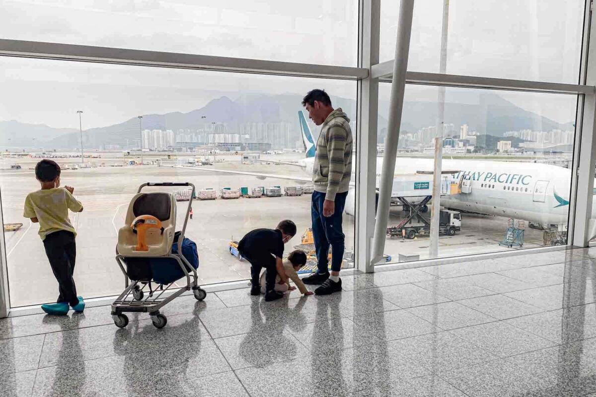 Dad stands with three young kids by Hong Kong airport windows watching Cathay Pacific planes before flying long haul.