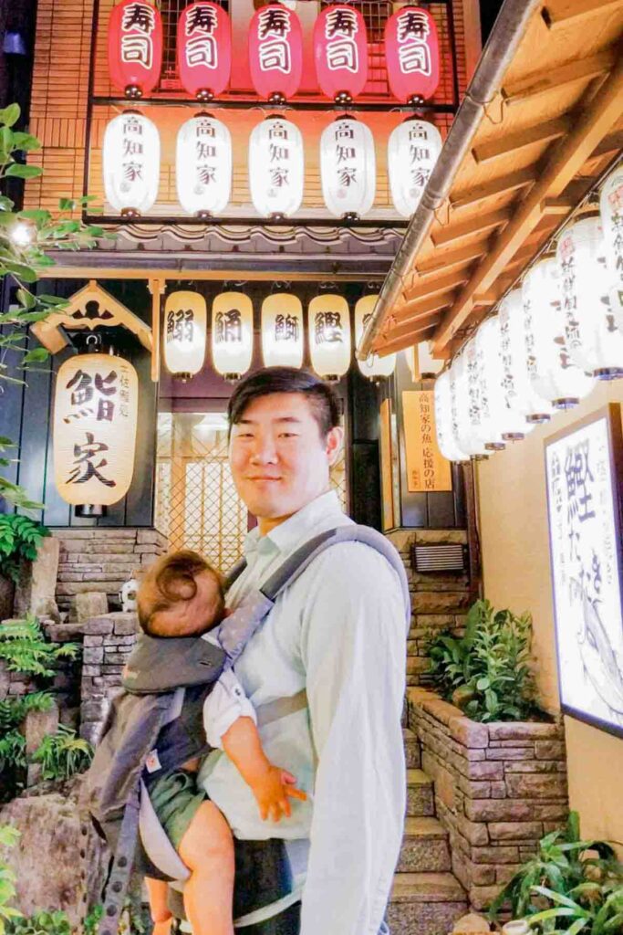 Father wearing a baby in a front carrier standing beneath rows of glowing restaurant lanterns in Osaka. Even evening food strolls work in Japan with a baby.