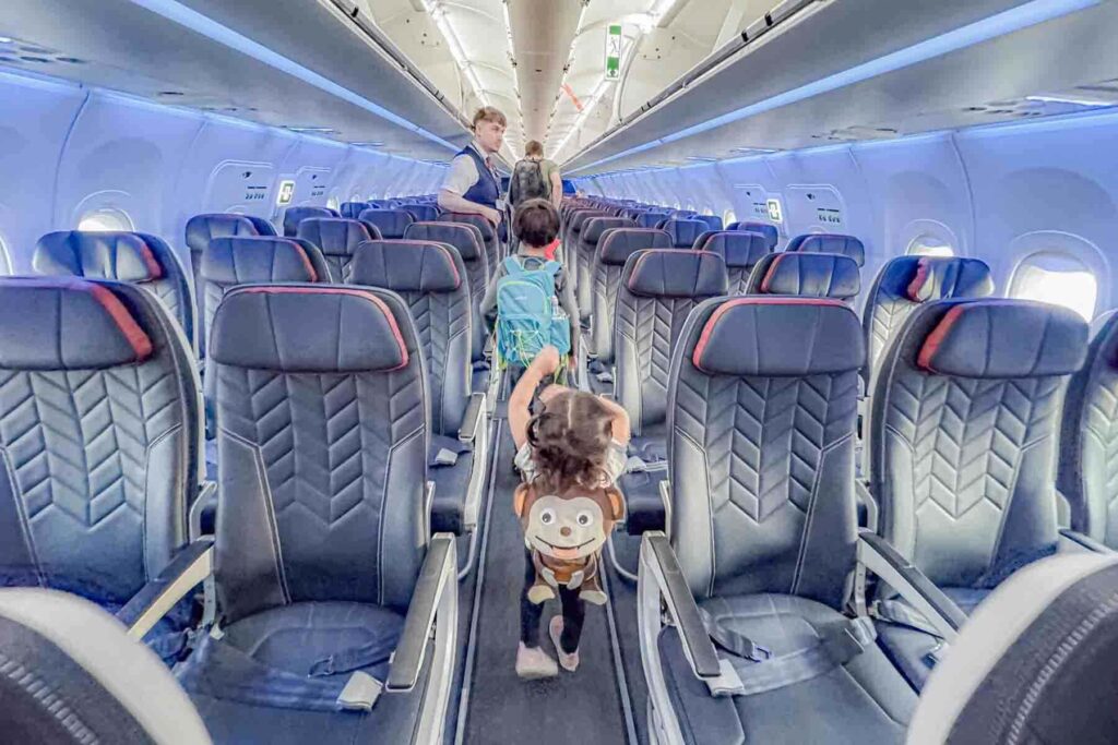 A toddler wearing a monkey backpack walks down the airplane aisle with her arms raised in triumph toward a flight attendant during deplaning after a flight. She follows her siblings through the empty cabin.
