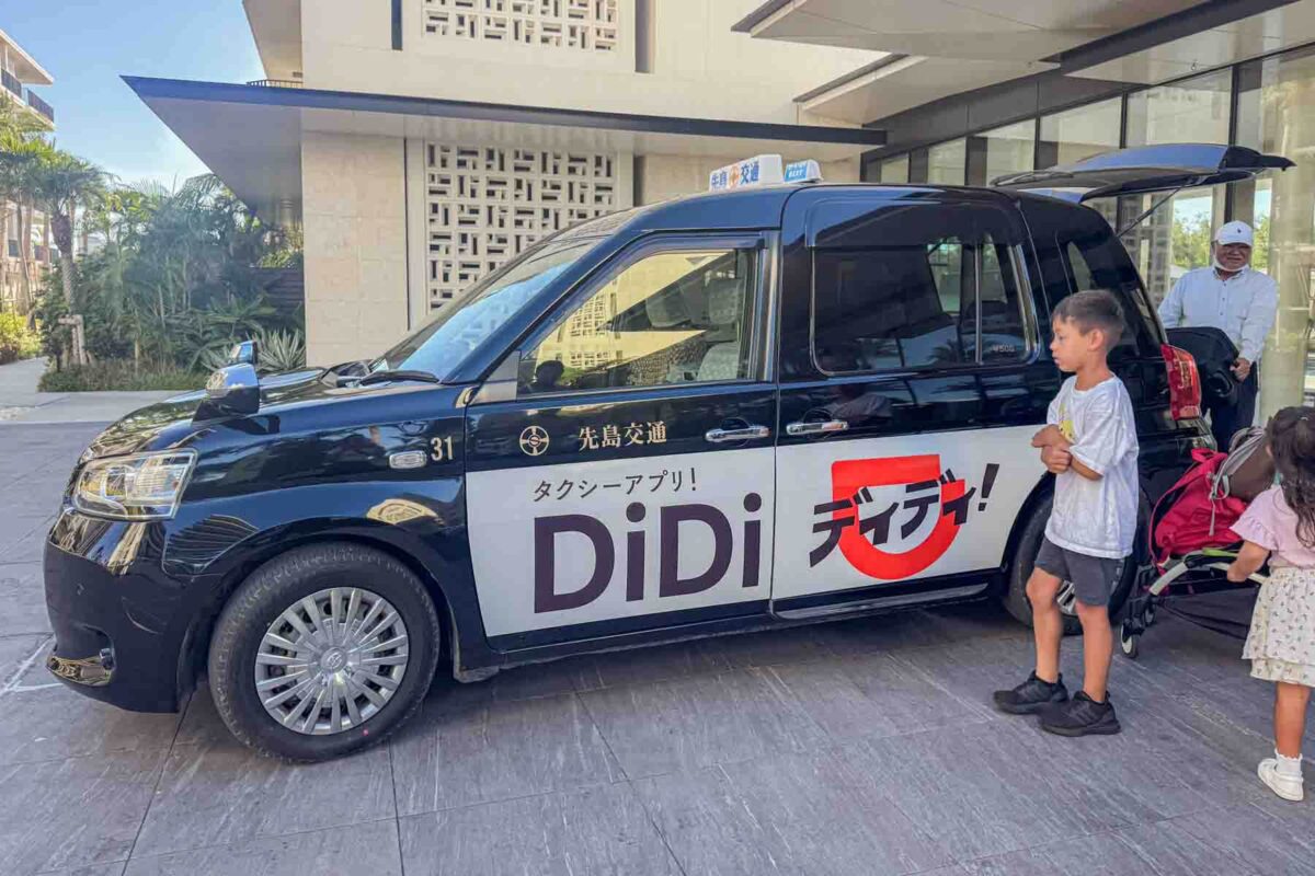 Family loading into a DiDi taxi outside a hotel in Okinawa with a stroller and luggage visible. A convenient option for getting around Japan with kids when public transit is less practical.