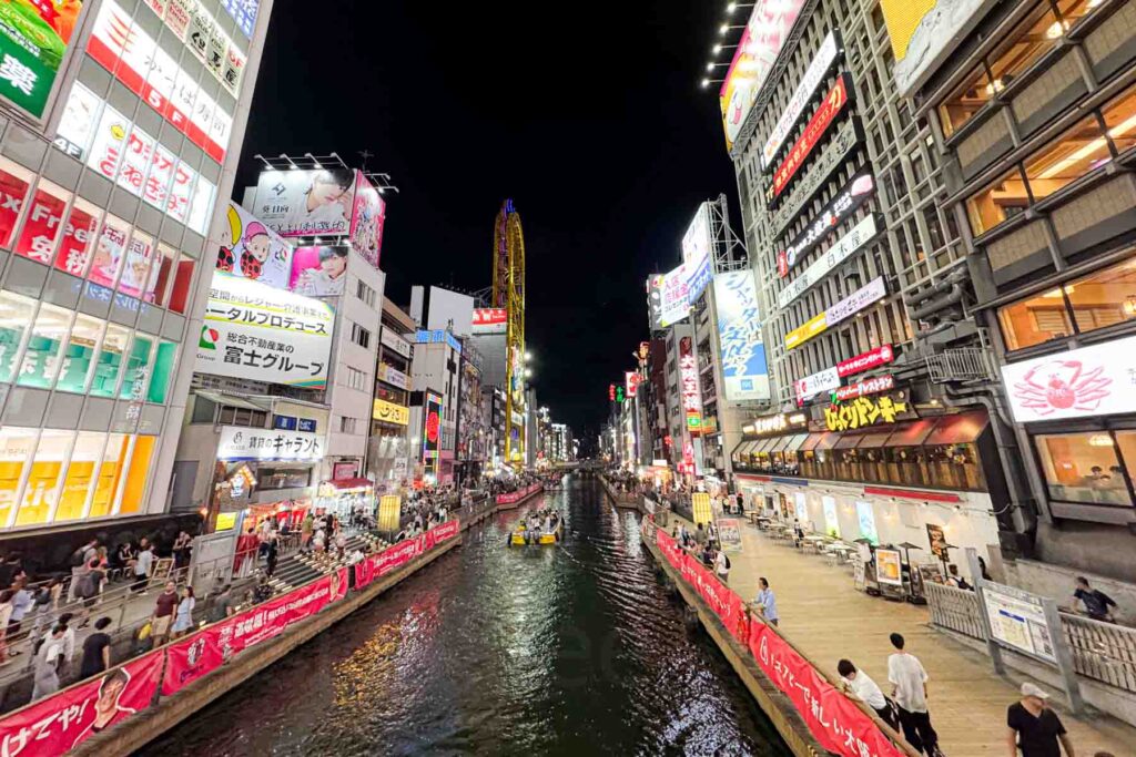 Night view of the Dotonbori canal in Osaka from the Ebisu Bridge lined with neon signs restaurants and crowds. This lively district shows the bright city side of exploring Japan with kids.