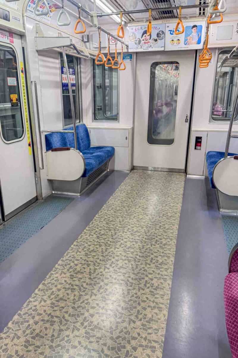 Empty Japanese metro car with blue seats and orange handle straps showing wide open floor space. A quieter moment outside of peak hours.