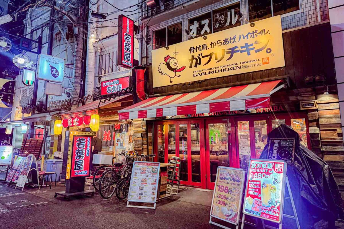 Neon lit street in Osaka with a casual Gaburi Chicken restaurant featuring striped awnings, bright signage, and menu boards outside. A lively place to eat in Japan.