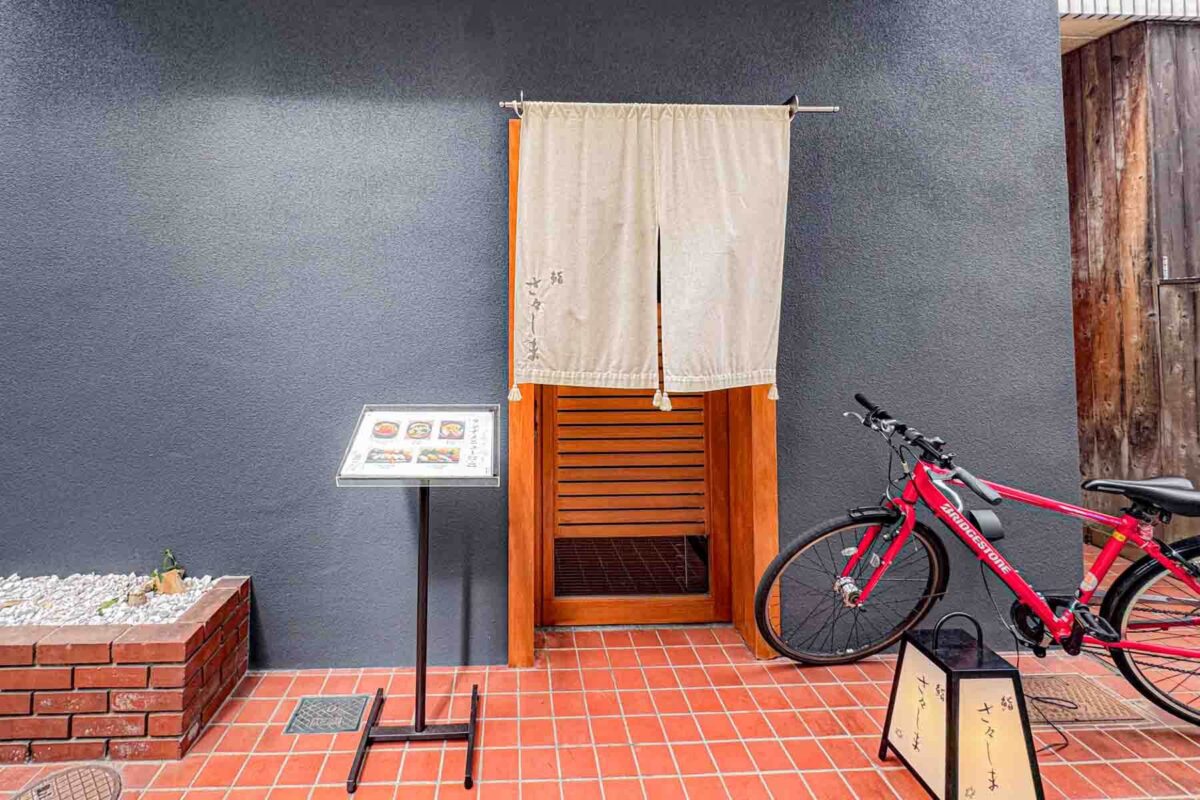 Minimalist restaurant entrance with a noren curtain, a standing menu sign, and a red bicycle nearby. A quiet neighborhood sushi spot for eating in Japan.