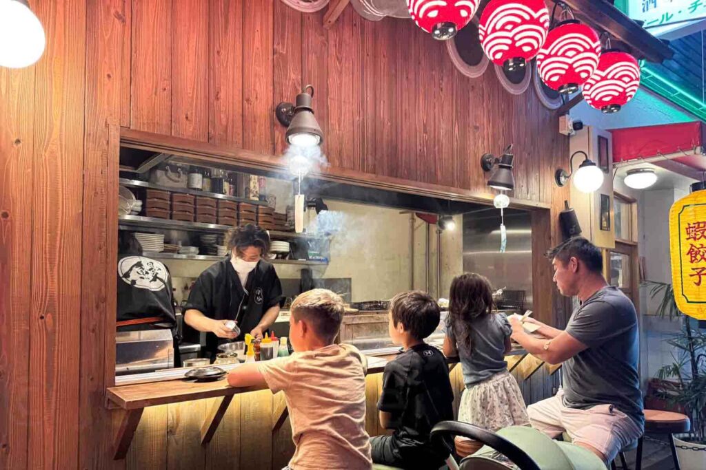Family with children sitting at a small street food counter with red lanterns hanging above them in Osaka while a chef cooks yakitori at the grill right in front of them. Casual local meals like this are fun when traveling to Japan with kids.