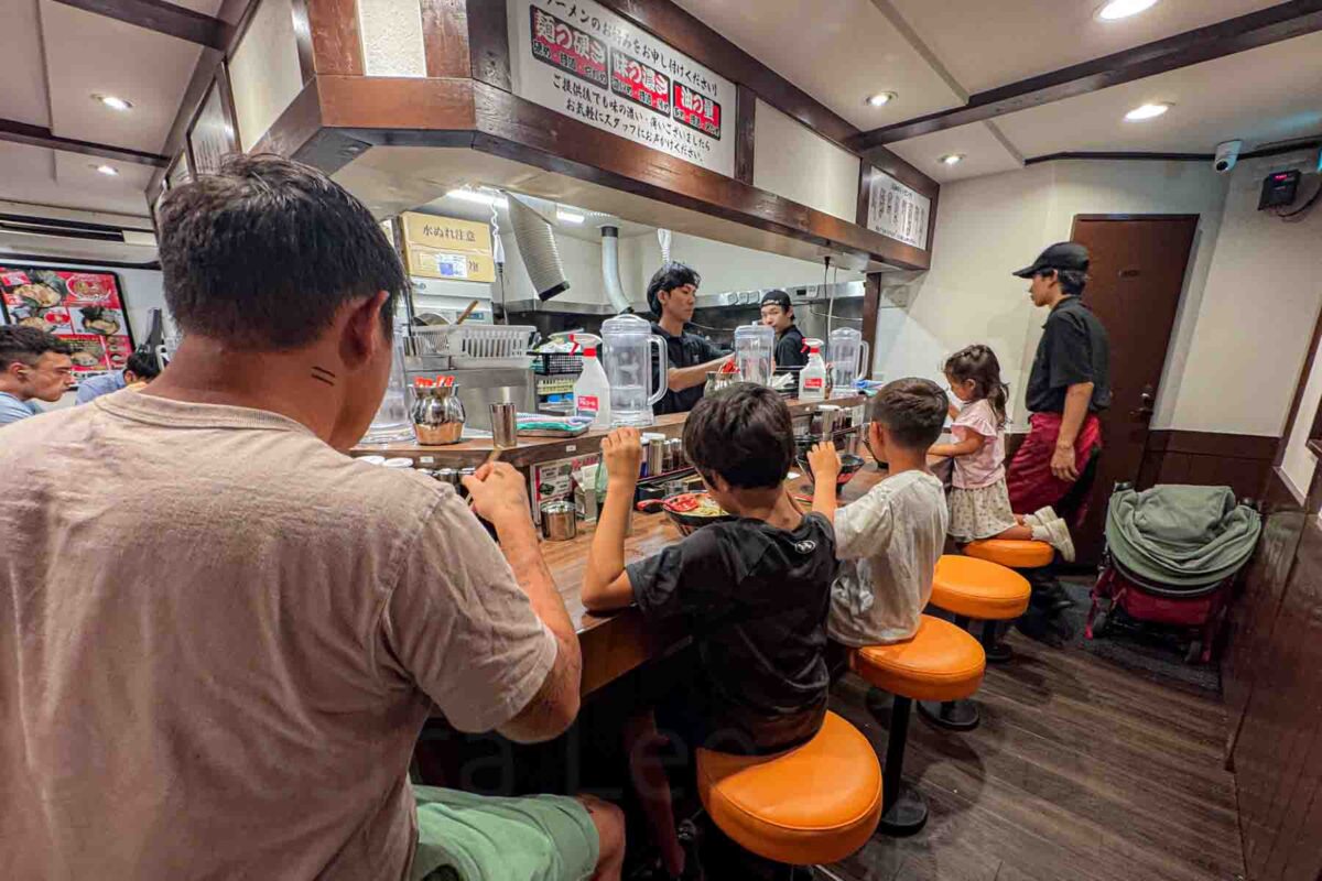 Family with children seated at a counter in a casual Japanese ramen restaurant in Tokyo watching chefs prepare food in an open kitchen. The kids sit on orange stools eating while staff cook behind the bar, showing a real experience of eating in Japan with kids in a lively local setting.
