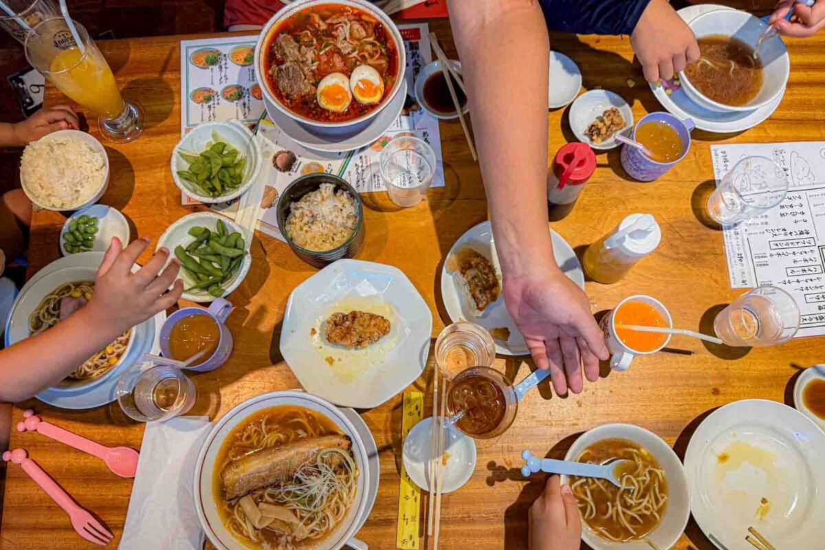 Overhead view of a table filled with ramen bowls, rice, edamame, and drinks as hands reach in to eat. A shared meal that reflects eating as a family of 5 in Japan.