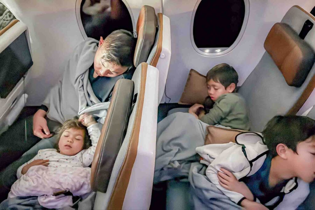 Three children and a parent sleep across two rows of airplane seats with pillows and blankets beside the window seats. The quiet scene shows how families manage sleep on a long haul flight to Asia.