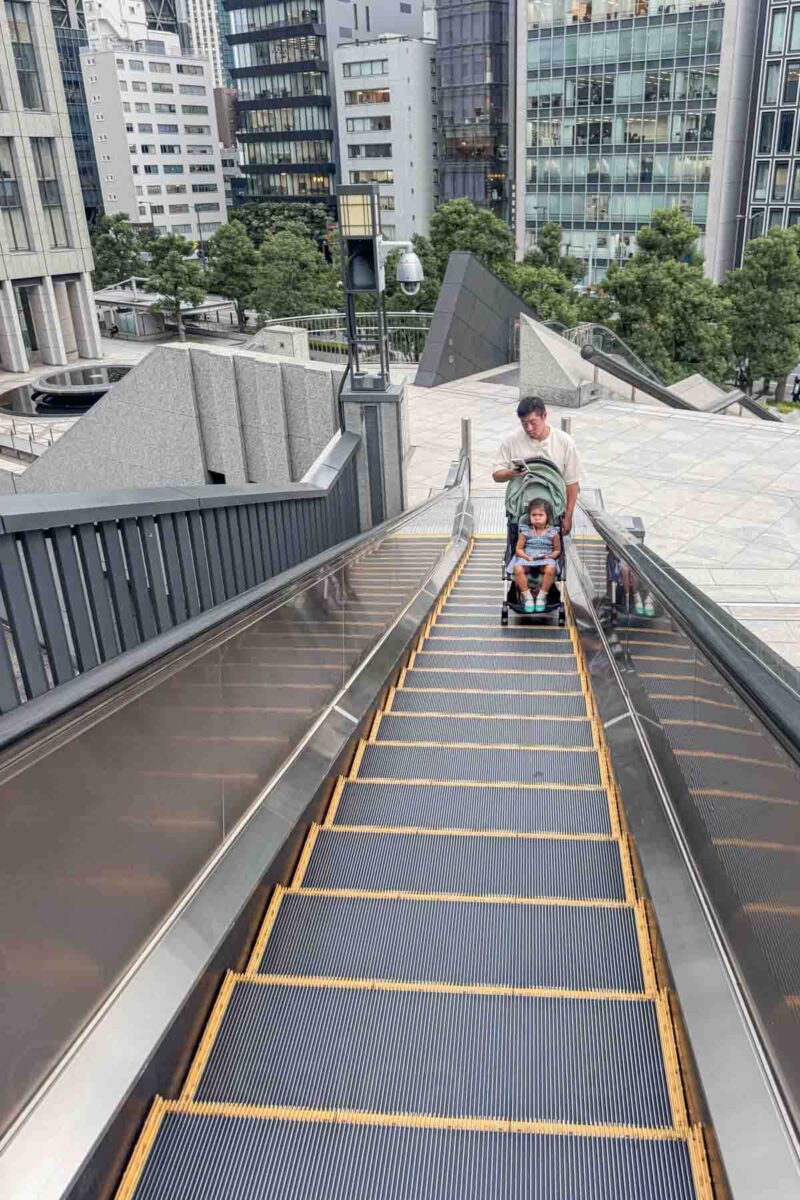 Parent holding a stroller up a long outdoor escalator in a modern Tokyo city area surrounded by tall buildings. Shows accessibility options helpful for Japan stroller travel in urban spaces.