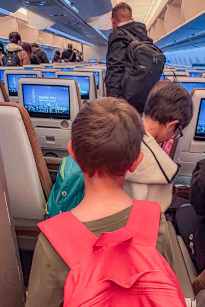 Two children wearing backpacks stand in the airplane aisle behind their father while passengers slowly exit the cabin. The scene shows the kids taking responsibility for their own belongings and the final moments of a long travel day with kids.