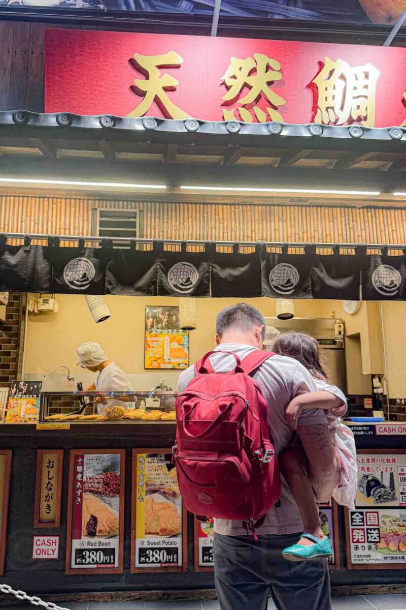 Adult wearing a red backpack holds a child while ordering at a taiyaki street food stall with signs reading “A Red Bean 380円”, “B Sweet Potato 380円”, and “C P 380円” with a “CASH ONLY” notice. The scene captures a quick and casual way of eating in Japan with kids at local food stands.