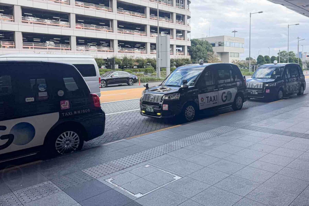 Line of GO taxis waiting at an airport pickup area in Japan with a parking structure in the background. Demonstrates taxis in Japan as a convenient airport transfer option.