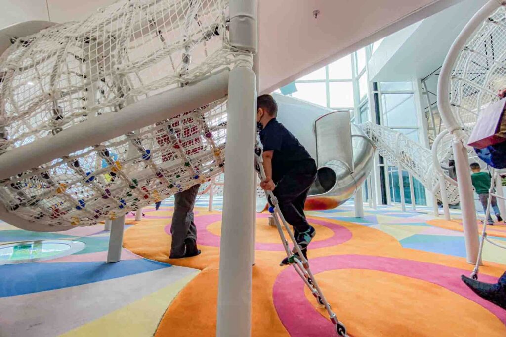Children climb white rope nets and slides above a soft clorful carpet area in an indoor play structure inside the Hong Kong airport terminal. Airport play areas help kids burn energy during long travel days.