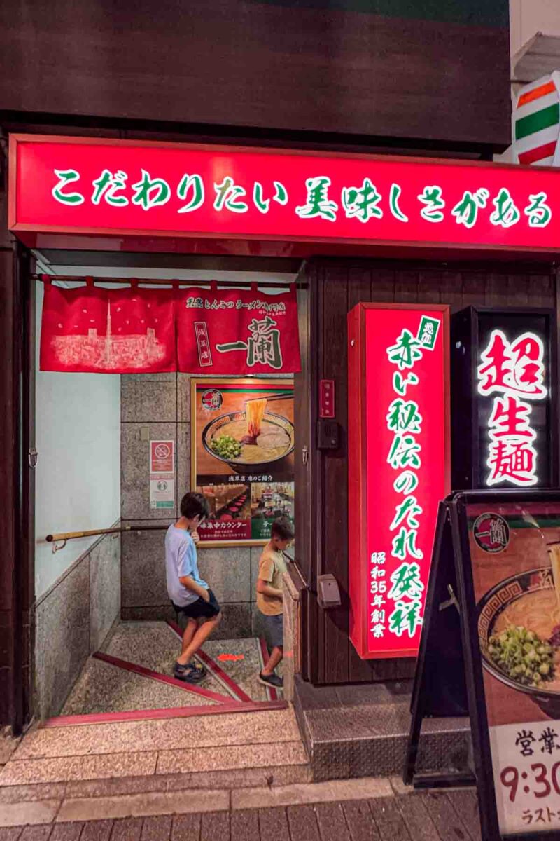 Red Ichiran restaurant entrance in Asakusa with Japanese signage and two children walking down stairs into a basement ramen shop. A unique dining setup that adds excitement for kids in Japan.
