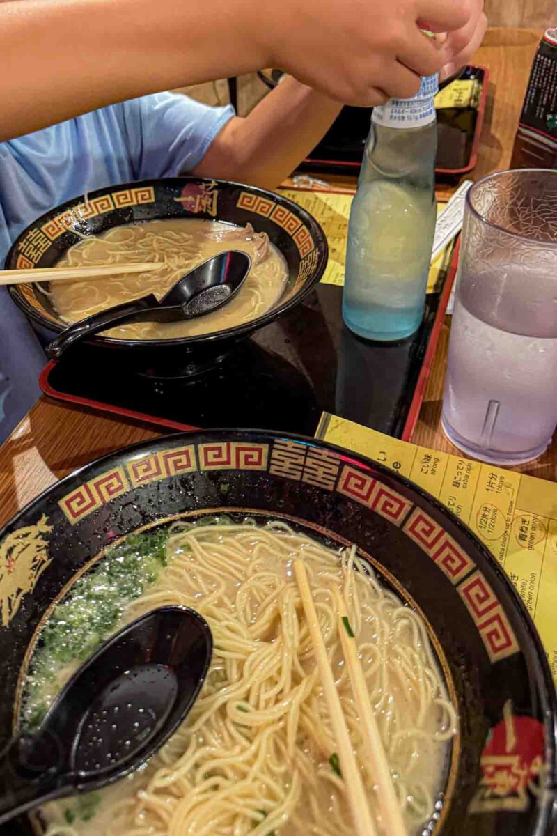 Two bowls of ramen with noodles and broth sit on a table at Ichiran Asakusa in Tokyo while a child opens a bottled drink beside a glass of water. The casual meal scene highlights a simple and easy option for families at a local restaurant.