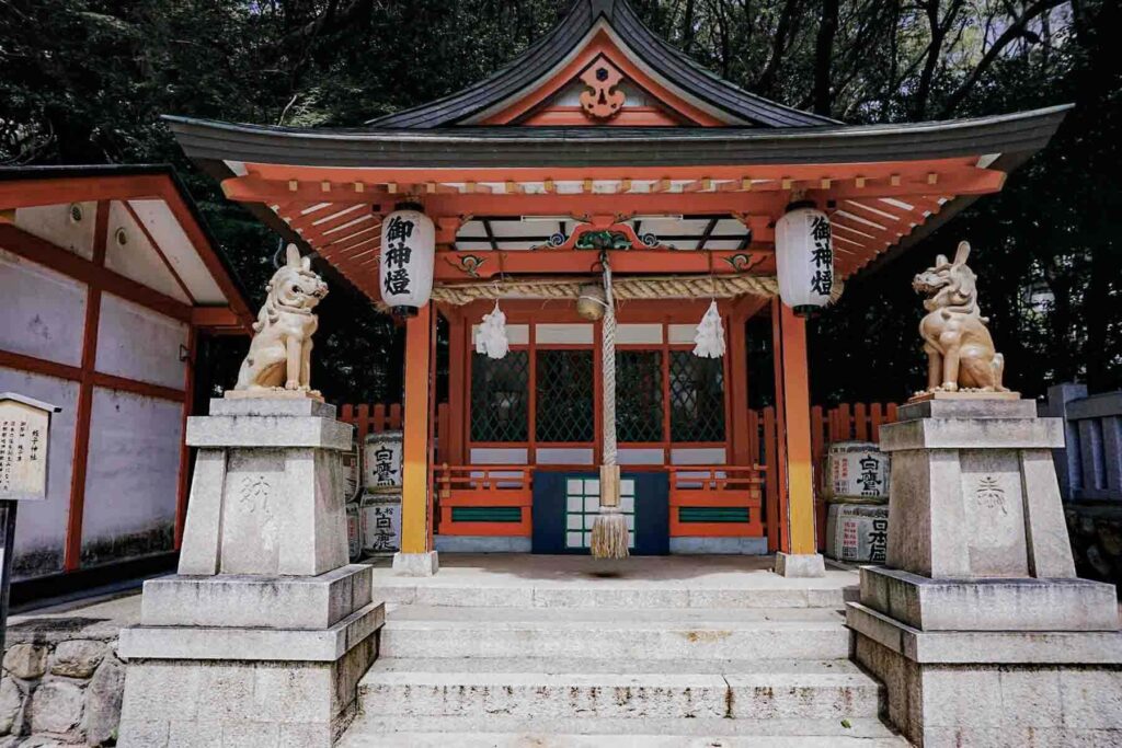 Small shrine building with orange beams and a curved roof at Ikuta Shrine in Kobe. Stone fox statues stand on both sides of the entrance, with white lanterns and black Japanese characters hanging from both sides, and a large rope hanging down in the middle attached to a bell.