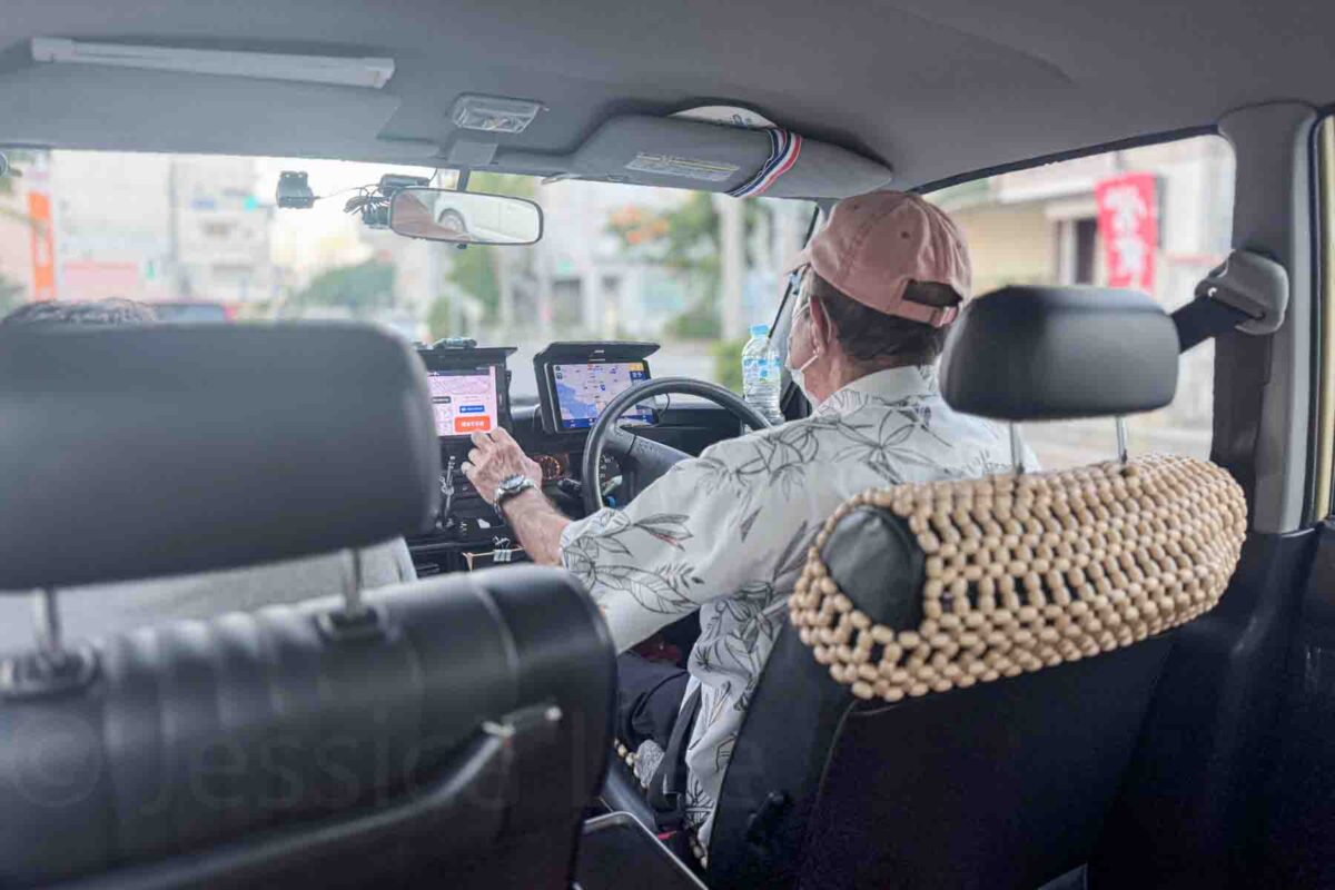 View from the back seat of a taxi in Japan with a driver in a baseball cap and white Hawaiian shirt navigating using a digital screen and dashboard controls. Highlights taxis in Okinawa as a stress free transport option.