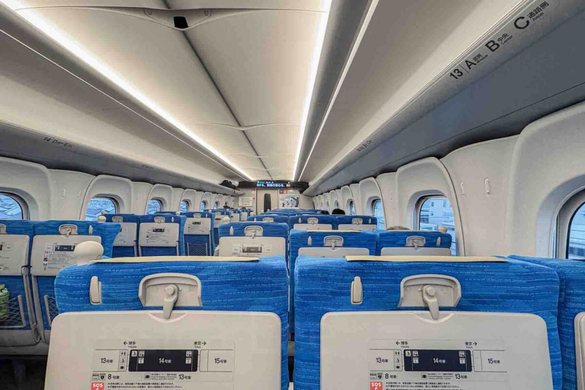Interior of a Shinkansen train with rows of blue seats and tray tables facing forward. Comfortable seating makes long distance travel easier when traveling around Japan with kids.
