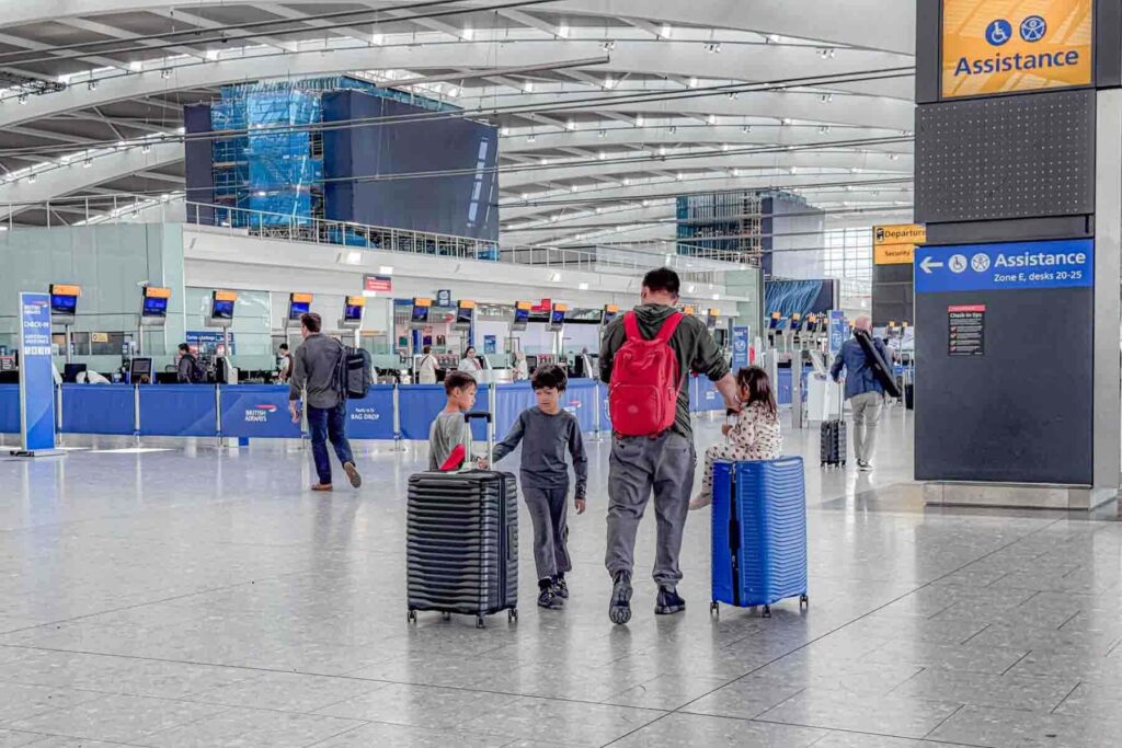A father walks wearing a red backpack through London Heathrow Airport with three children pulling rolling suitcases past check in counters. A sign reads "Assistance Zone E desks 20-25".